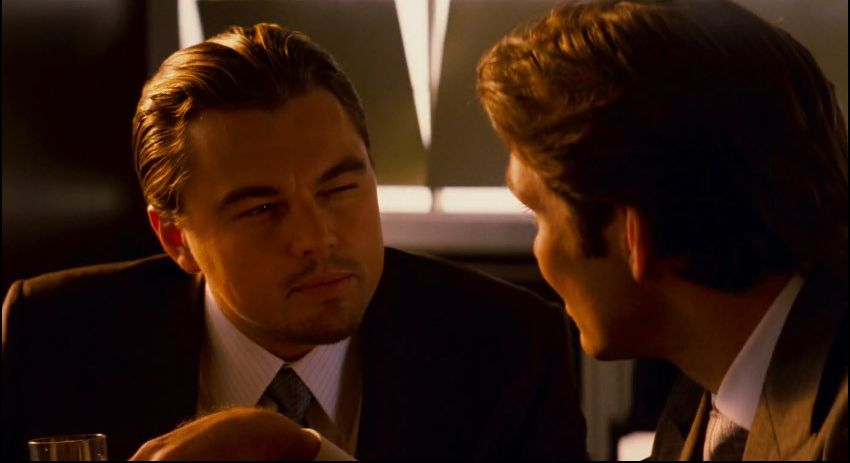 Still from Inception of two characters talking across a table in a dim bar